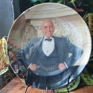 May include: A ceramic plate with a portrait of a man in a suit and bow tie. The plate is on a black stand. The man is smiling and has a bald head. The background is a light beige color.
