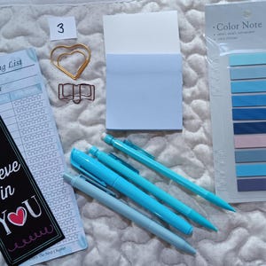 May include: A reading list with a checklist, a bookmark that says "I believe in you", a blue sticky note, a gold paper clip shaped like a book, a gold paper clip shaped like two hearts, and four blue pens on a white quilted surface.