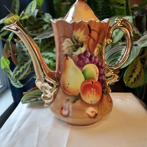 May include: A brown and gold ceramic teapot with a fruit design. The teapot has a lid with a gold knob and a handle with a gold trim. The fruit design includes pears, grapes, and apples.