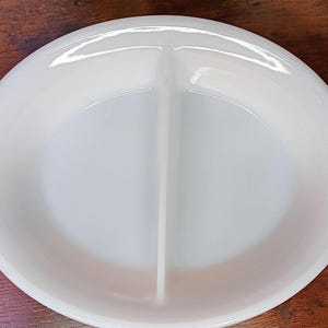 May include: A white, oval-shaped divided serving dish with two sections. The dish has rounded edges and two handles. The dish is made of a glossy, opaque material. The dish is on a wooden surface.