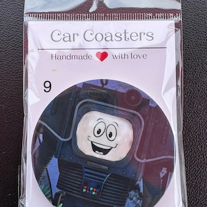 May include: A round car coaster featuring a cartoon robot with a smiling face and a television screen for a head. The robot is black and gray with a blue and green screen. The coaster is made of a durable material and is perfect for protecting your car's cup holders from spills and scratches.