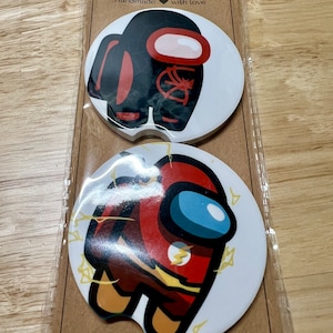 May include: Two round car coasters featuring the Among Us character in different superhero costumes. The top coaster features the character dressed as Spider-Man, and the bottom coaster features the character dressed as The Flash. The coasters are white with a brown background and the text "Car Coasters" and "Handmade with love".
