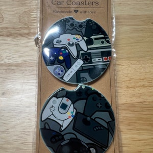 May include: Two car coasters with a gaming theme. The coasters are round and feature a collage of gray and white game controllers and consoles. The packaging reads "Car Coasters" and "Handmade with love."