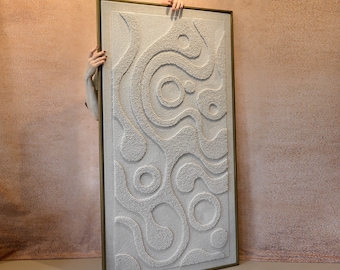May include: A rectangular, off-white textured wall art piece with a gold frame. The artwork features a raised, swirling pattern of circles and lines. The piece is held by two hands against a peach-colored background.