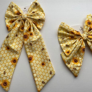 May include: Two yellow and brown fabric hair bows with a honeycomb and sunflower pattern. The larger bow is a long, single bow, while the smaller bow is a traditional double bow.
