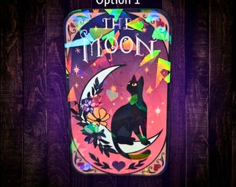 May include: A rectangular sticker featuring a black cat sitting on a crescent moon with the text THE MOON above. The design includes colourful floral accents, small stars, and a heart at the bottom, all finished with a holographic overlay effect.