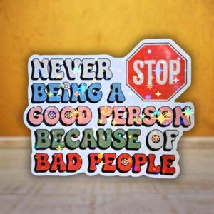 Puede incluir: Una pegatina holográfica con una señal de stop octogonal roja y el texto NEVER BEING A GOOD PERSON BECAUSE OF BAD PEOPLE. Las letras utilizan una fuente retro multicolor en azul, rosa, verde y rojo, acentuadas por destellos en forma de estrella.