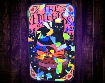 May include: A rectangular sticker featuring a black cat with small white wings sitting on a stack of pillows. The text THE EMPRESS is printed at the top. The design includes a pink background with heart and star accents, overlaid with iridescent shards.