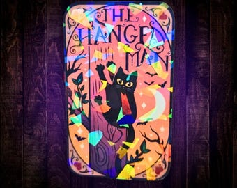 May include: A rectangular sticker with a holographic finish featuring a black cat climbing a tree. The text THE HANGED MAN is printed at the top. The background is a warm orange hue with illustrations of a crescent moon, small bats, and branches.