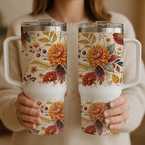 Könnte beinhalten: Zwei weiße, isolierte Becher mit floralen Mustern. Die Becher haben einen cremefarbenen Hintergrund mit orangefarbenen, roten und gelben Blumen und Blättern. Jeder Becher hat einen weißen Griff und einen silbernen Deckel.