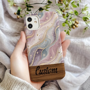 Op de afbeelding: Een op maat gemaakte telefoonhoes met een wervelend sterrenstelselontwerp in paars, blauw en goud. De hoes heeft een houten onderkant met het woord "Custom" gegraveerd. Ontworpen voor een smartphone.