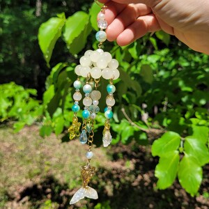 May include: A hanging ornament featuring a cluster of translucent off-white beads at the top, with dangling strands of turquoise, white, and clear beads. The ornament includes small yellow star and clear shell charms, finished with a gold glitter mermaid tail.