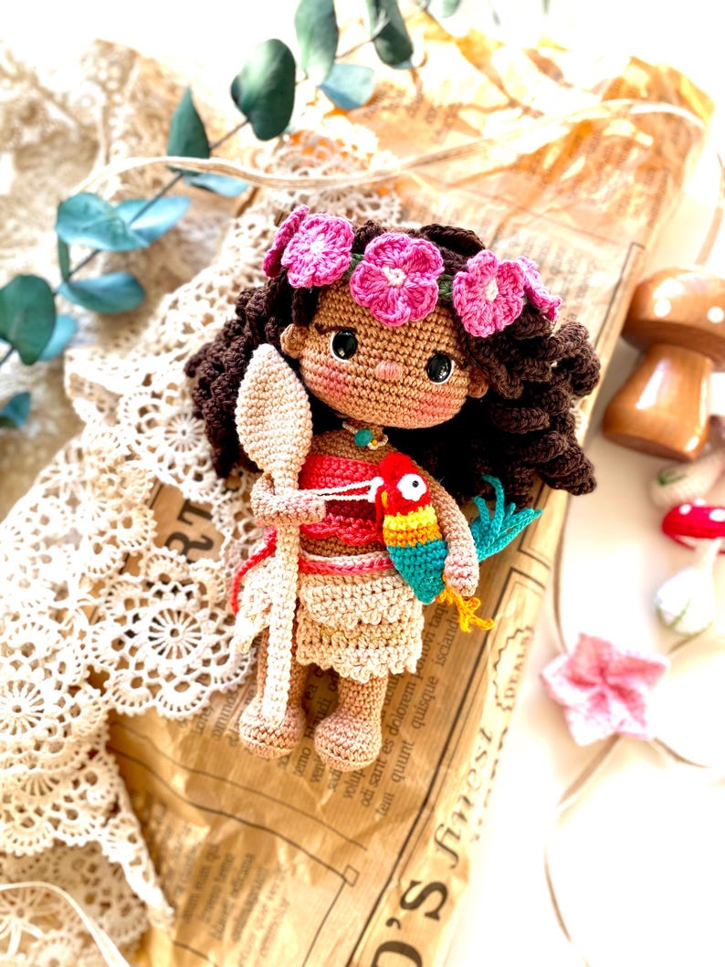 Puede incluir: Mu&ntilde;eca de ganchillo hecha a mano con cabello rizado casta&ntilde;o oscuro, una corona de flores rosas y una parte superior roja. La mu&ntilde;eca sostiene una cuchara de madera y un loro colorido. La mu&ntilde;eca lleva una falda beige y un collar turquesa.