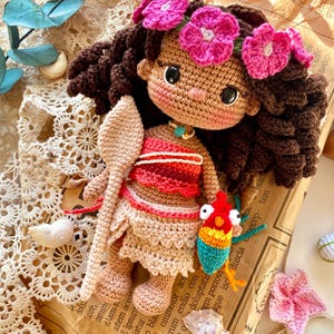 Puede incluir: Mu&ntilde;eca de ganchillo hecha a mano con cabello rizado casta&ntilde;o, una corona de flores rosas y un atuendo rojo y beige. La mu&ntilde;eca sostiene una cuchara de madera y un peque&ntilde;o pez colorido. Sobre un fondo de papel vintage.
