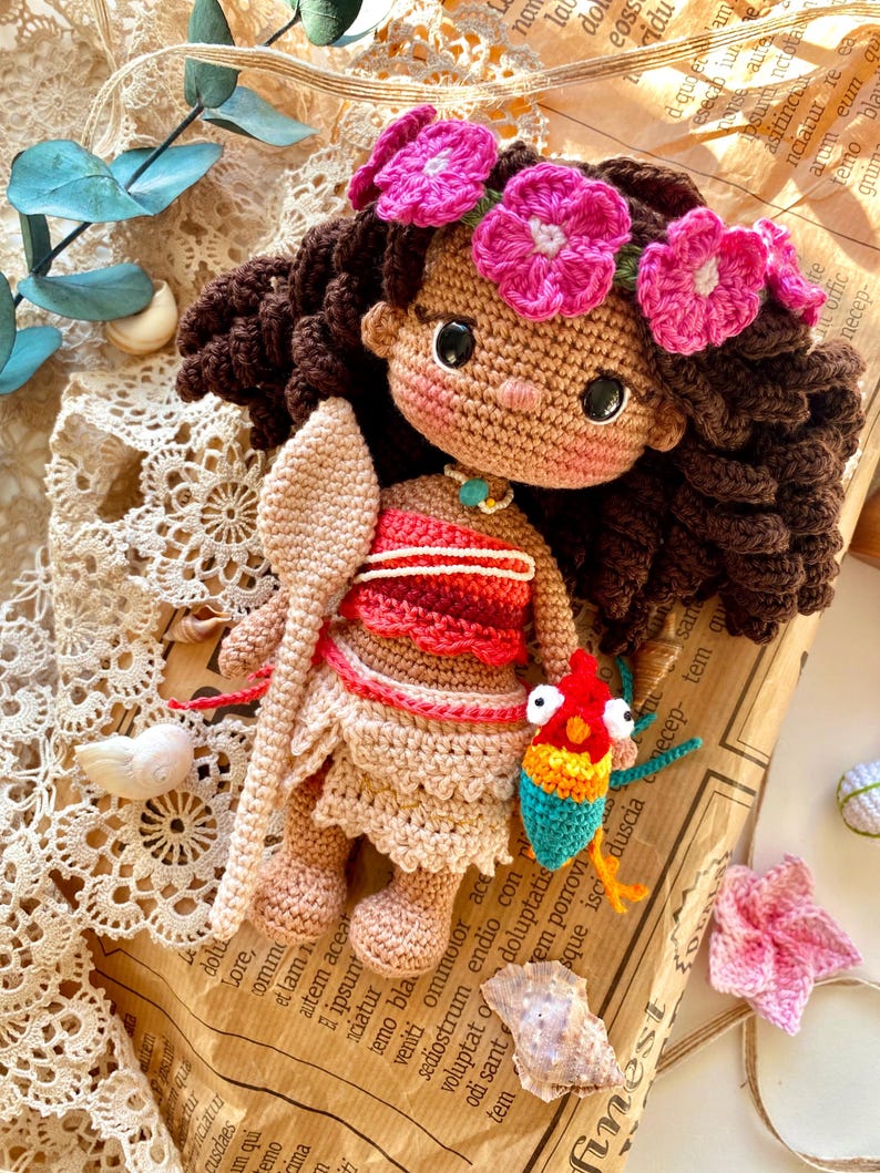 Puede incluir: Mu&ntilde;eca de ganchillo hecha a mano con cabello rizado casta&ntilde;o, una corona de flores rosas y un atuendo rojo y beige. La mu&ntilde;eca sostiene una cuchara de madera y un peque&ntilde;o pez colorido. Sobre un fondo de papel vintage.