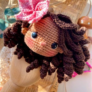 Puede incluir: Cabeza de mu&ntilde;eca de ganchillo hecha a mano con cabello rizado marr&oacute;n y una flor rosa en la parte superior. La mu&ntilde;eca tiene ojos de bot&oacute;n negros y mejillas rosadas. La mu&ntilde;eca est&aacute; hecha de hilo y es un art&iacute;culo artesanal.