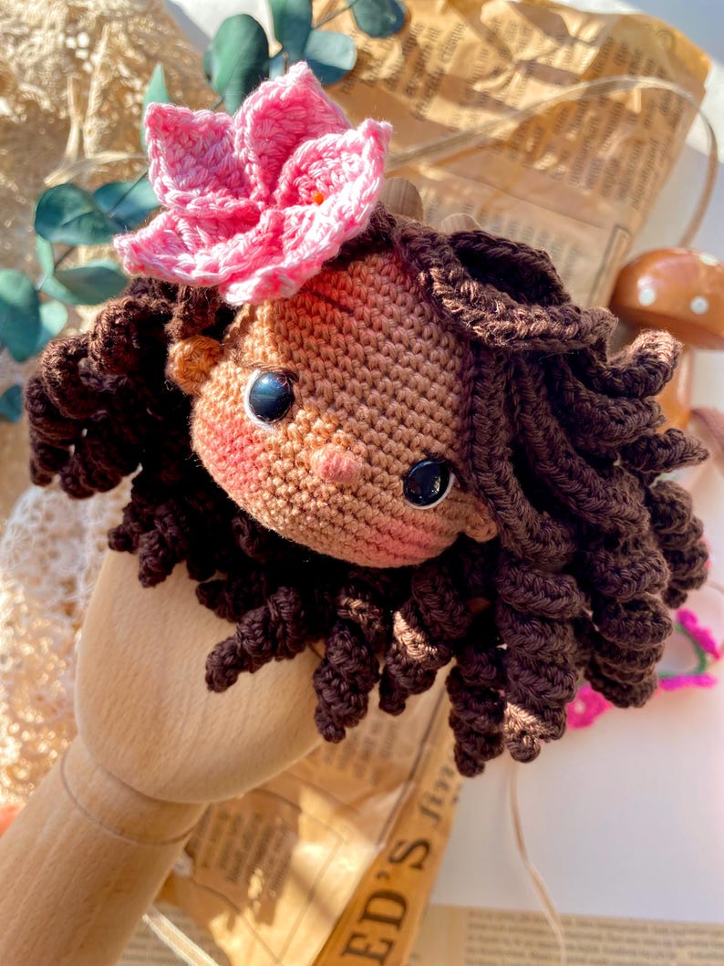 Puede incluir: Cabeza de mu&ntilde;eca de ganchillo hecha a mano con cabello rizado marr&oacute;n y una flor rosa en la parte superior. La mu&ntilde;eca tiene ojos de bot&oacute;n negros y mejillas rosadas. La mu&ntilde;eca est&aacute; hecha de hilo y es un art&iacute;culo artesanal.
