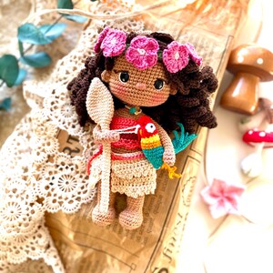 Puede incluir: Mu&ntilde;eca de ganchillo hecha a mano con cabello casta&ntilde;o, una corona de flores rosas y un atuendo rojo y beige. La mu&ntilde;eca sostiene una cuchara de madera y un loro colorido. La mu&ntilde;eca est&aacute; sobre un fondo de papel vintage.