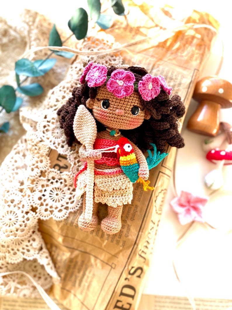 Puede incluir: Mu&ntilde;eca de ganchillo hecha a mano con cabello casta&ntilde;o, una corona de flores rosas y un atuendo rojo y beige. La mu&ntilde;eca sostiene una cuchara de madera y un loro colorido. La mu&ntilde;eca est&aacute; sobre un fondo de papel vintage.