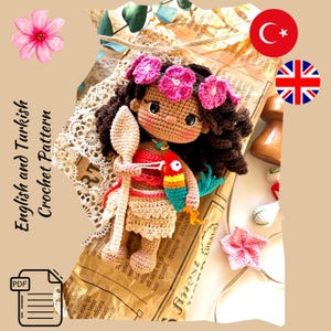 Puede incluir: Una mu&ntilde;eca de ganchillo hecha a mano con cabello casta&ntilde;o oscuro, vestida con un atuendo rojo y beige, y una corona de flores rosas. La mu&ntilde;eca sostiene una cuchara de madera y un loro colorido. El texto "English and Turkish Crochet Pattern" est&aacute; incluido.
