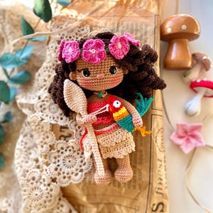 Puede incluir: Mu&ntilde;eca de ganchillo con cabello casta&ntilde;o oscuro, una corona de flores rosas y un atuendo rojo y beige. La mu&ntilde;eca sostiene una cuchara de madera y un loro colorido. Hongos de madera y una flor rosa en el fondo.