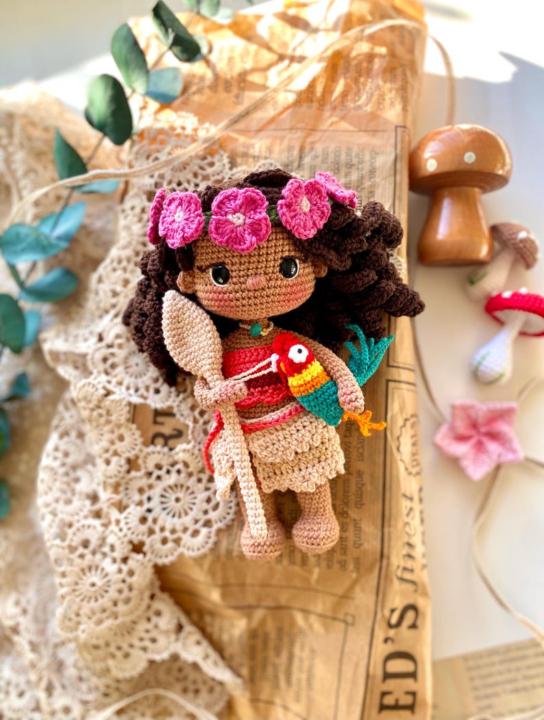 Puede incluir: Mu&ntilde;eca de ganchillo con cabello casta&ntilde;o oscuro, una corona de flores rosas y un atuendo rojo y beige. La mu&ntilde;eca sostiene una cuchara de madera y un loro colorido. Hongos de madera y una flor rosa en el fondo.