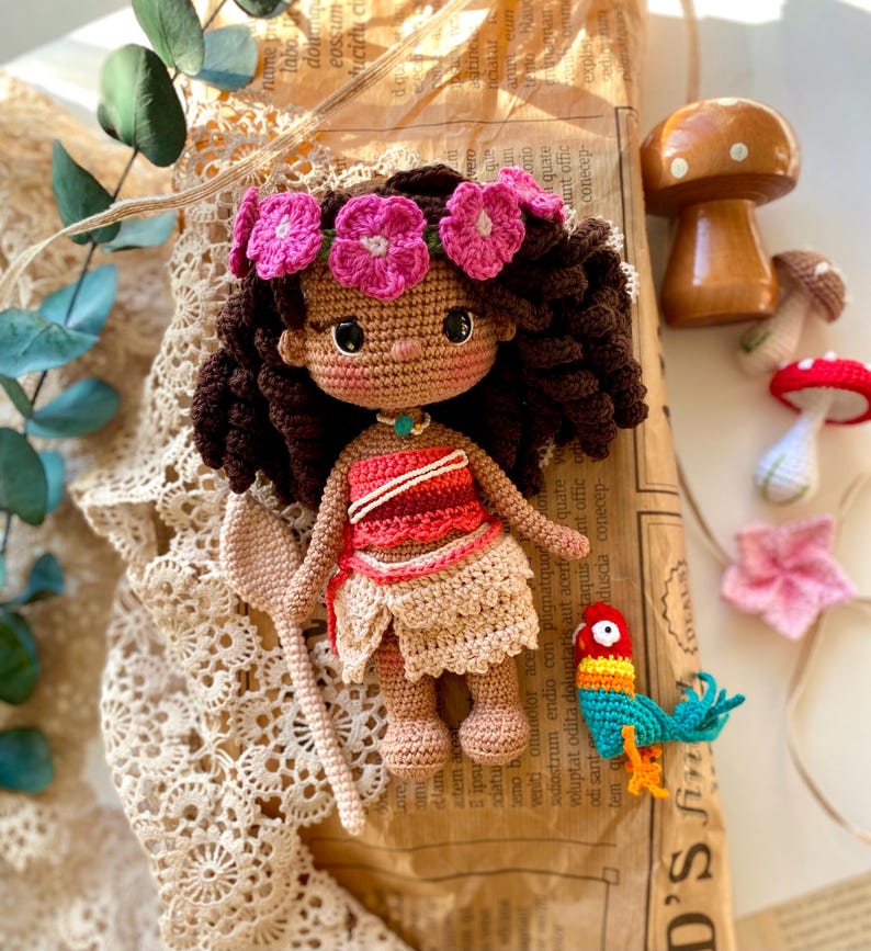 Puede incluir: Mu&ntilde;eca de ganchillo hecha a mano con cabello casta&ntilde;o, una corona de flores rosas y una parte superior roja y blanca. La mu&ntilde;eca sostiene una cuchara de ganchillo y est&aacute; acompa&ntilde;ada de un loro de ganchillo y decoraciones de hongos.