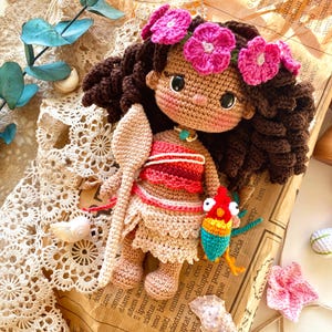 Puede incluir: Mu&ntilde;eca de ganchillo con cabello rizado marr&oacute;n oscuro, vestida con un atuendo rojo y beige y una corona de flores rosas. La mu&ntilde;eca sostiene una cuchara de madera y un p&aacute;jaro colorido. La mu&ntilde;eca est&aacute; sobre papel vintage.