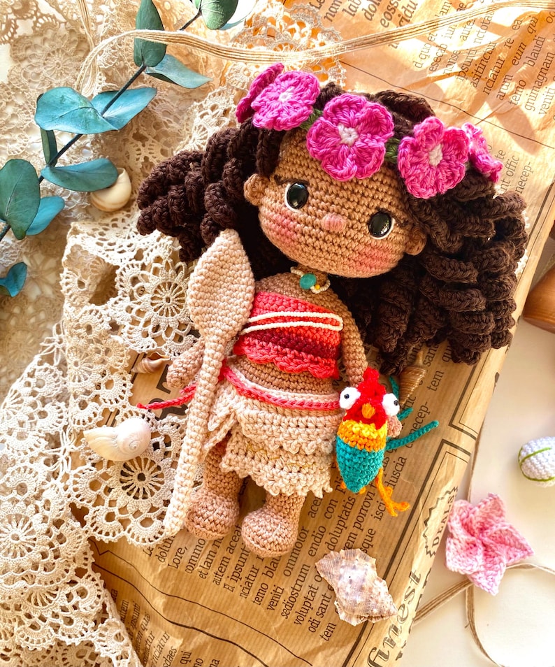 Puede incluir: Mu&ntilde;eca de ganchillo con cabello rizado marr&oacute;n oscuro, vestida con un atuendo rojo y beige y una corona de flores rosas. La mu&ntilde;eca sostiene una cuchara de madera y un p&aacute;jaro colorido. La mu&ntilde;eca est&aacute; sobre papel vintage.
