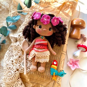 Puede incluir: Una mu&ntilde;eca de ganchillo hecha a mano con cabello casta&ntilde;o, vestida con un atuendo rojo y beige, y adornada con una corona de flores rosas. La mu&ntilde;eca sostiene una paleta de ganchillo y est&aacute; junto a un loro de ganchillo colorido. Tambi&eacute;n hay una seta de madera y otros art&iacute;culos de ganchillo.