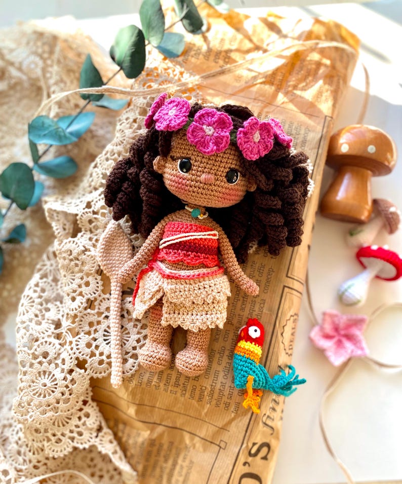 Puede incluir: Una mu&ntilde;eca de ganchillo hecha a mano con cabello casta&ntilde;o, vestida con un atuendo rojo y beige, y adornada con una corona de flores rosas. La mu&ntilde;eca sostiene una paleta de ganchillo y est&aacute; junto a un loro de ganchillo colorido. Tambi&eacute;n hay una seta de madera y otros art&iacute;culos de ganchillo.