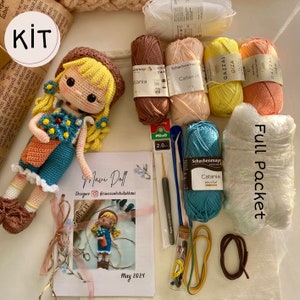 May include: A crochet doll kit with yarn, crochet hooks, and a pattern. The kit includes a variety of colors of yarn, including peach, yellow, brown, and blue. The doll is wearing a blue and orange dress with a flower pattern. The kit also includes a small glass bottle with beads and a brown cord.