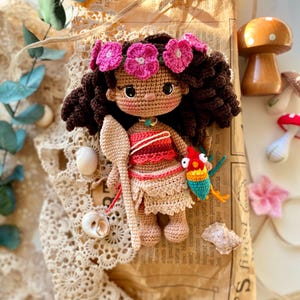 Puede incluir: Mu&ntilde;eca de ganchillo hecha a mano con cabello rizado casta&ntilde;o oscuro, una corona de flores rosas y un atuendo rojo y beige. La mu&ntilde;eca sostiene una cuchara de madera y un p&aacute;jaro colorido. Cerca hay conchas marinas decorativas y una seta de madera.