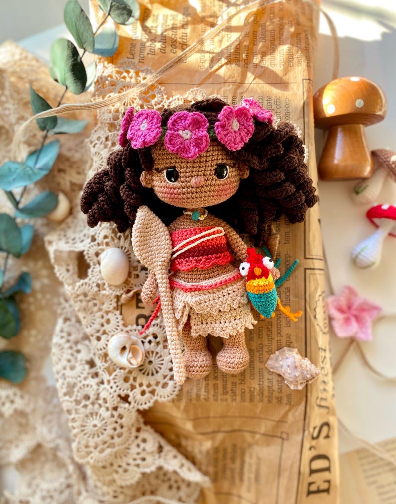 Puede incluir: Mu&ntilde;eca de ganchillo hecha a mano con cabello rizado casta&ntilde;o oscuro, una corona de flores rosas y un atuendo rojo y beige. La mu&ntilde;eca sostiene una cuchara de madera y un p&aacute;jaro colorido. Cerca hay conchas marinas decorativas y una seta de madera.