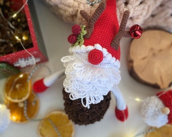 Op de afbeelding: Een gehaakt rendier ornament met een rode muts, witte baard, bruine geweien en een belletje. Het ornament is omringd door gedroogde sinaasappelschijfjes en een lichtsnoer.