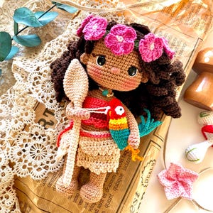 Puede incluir: Una mu&ntilde;eca de ganchillo hecha a mano con cabello rizado casta&ntilde;o, una corona de flores rosas y tez bronceada. La mu&ntilde;eca viste un atuendo rojo y beige y sostiene una cuchara de ganchillo y un loro colorido. La mu&ntilde;eca est&aacute; colocada sobre un fondo de papel vintage.