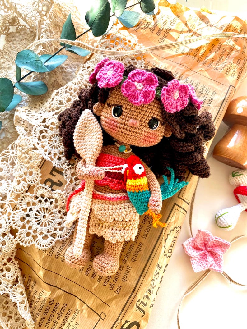 Puede incluir: Una mu&ntilde;eca de ganchillo hecha a mano con cabello rizado casta&ntilde;o, una corona de flores rosas y tez bronceada. La mu&ntilde;eca viste un atuendo rojo y beige y sostiene una cuchara de ganchillo y un loro colorido. La mu&ntilde;eca est&aacute; colocada sobre un fondo de papel vintage.