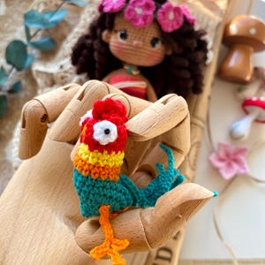 Puede incluir: Un loro de ganchillo hecho a mano y colorido con detalles rojos, amarillos, naranjas y turquesas. Una mu&ntilde;eca de ganchillo con una corona de flores rosas est&aacute; en el fondo. El loro se sostiene en una mano de madera.