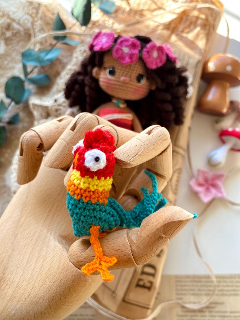 Puede incluir: Un loro de ganchillo hecho a mano y colorido con detalles rojos, amarillos, naranjas y turquesas. Una mu&ntilde;eca de ganchillo con una corona de flores rosas est&aacute; en el fondo. El loro se sostiene en una mano de madera.