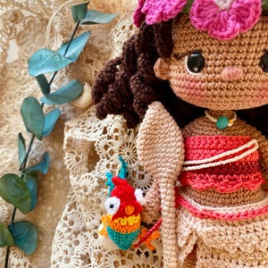 Puede incluir: Mu&ntilde;eca de ganchillo hecha a mano con cabello casta&ntilde;o, una corona de flores rosas y una parte superior roja y blanca. La mu&ntilde;eca sostiene una lanza de ganchillo y un peque&ntilde;o pez colorido. La mu&ntilde;eca tiene piel bronceada y un collar con un colgante turquesa.