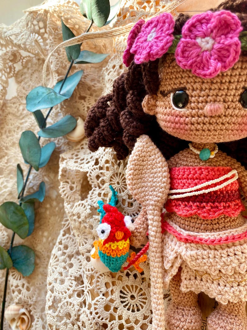 Puede incluir: Mu&ntilde;eca de ganchillo hecha a mano con cabello casta&ntilde;o, una corona de flores rosas y una parte superior roja y blanca. La mu&ntilde;eca sostiene una lanza de ganchillo y un peque&ntilde;o pez colorido. La mu&ntilde;eca tiene piel bronceada y un collar con un colgante turquesa.