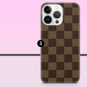 May include: A brown and tan checkered phone case with a clear border. The case has a camera cutout and the number 3 in a black circle. The background is a gradient of pink and white.