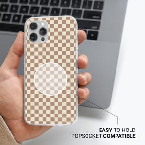 May include: Clear phone case with a brown and beige checkered pattern. The case has a popsocket-compatible design and is easy to hold. The phone case is held in a person's hand, with a laptop in the background.