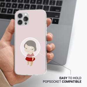 May include: A clear phone case with a light pink background featuring a cartoon illustration of a girl with an angry expression. The case is labeled "EASY TO HOLD POPSOCKET COMPATIBLE".