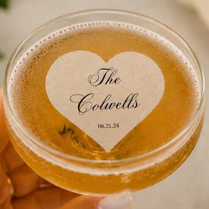 May include: A clear glass coupe filled with a golden liquid featuring a white heart-shaped garnish floating on top. The garnish is printed with the text The Colwells and the date 06.21.24 in a black serif font centered within the heart shape.