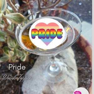 Puede incluir: Un corazón arcoíris con la palabra "PRIDE" en su interior, sobre una copa de martini de vidrio transparente. La copa está apoyada en una roca con las palabras "Pride Drink Toppers" debajo.