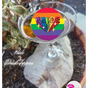 Peut inclure: Un accessoire de boisson arc-en-ciel avec le mot "PRIDE" en lettres blanches. L'accessoire est dans un verre à martini et repose sur un rocher.