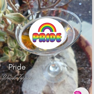 Può includere: Un adesivo rotondo bianco con un arcobaleno e la parola "PRIDE" in colori arcobaleno. L'adesivo è sopra un bicchiere da martini pieno di un liquido dorato. Il bicchiere è su uno scoglio.