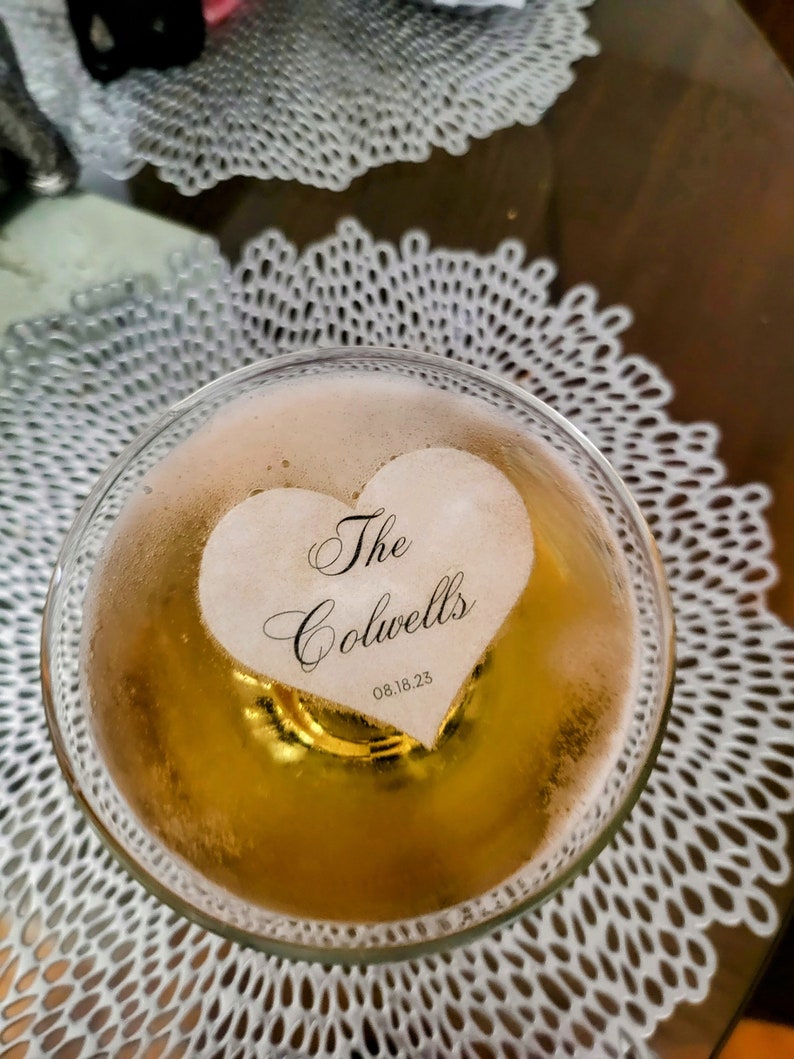 Peut inclure: Un gla&ccedil;on en forme de c&oelig;ur avec le texte "The Colwells 08.18.23" en calligraphie noire, flottant dans un verre de champagne.