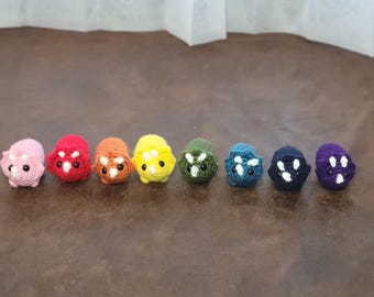 May include: A row of eight crocheted dinosaur toys in a rainbow of colors, including pink, red, orange, yellow, green, blue, teal, and purple.